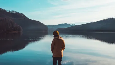 Person in Kapuzenpullover steht am Ufer eines spiegelglatten Bergsees