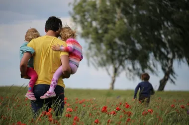 Ein Mann trägt zwei kleine Kinder auf den Armen durch ein Mohnblumenfeld, im Hintergrund läuft ein drittes Kind